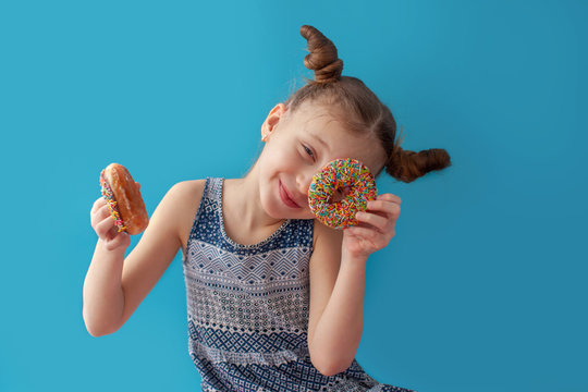 7 year old girl rejoices at the donut. She is in anticipation. She is enjoys. Hairstyle. On a blue background in a blue dress. Classic chocolate donut with sprinkles. 