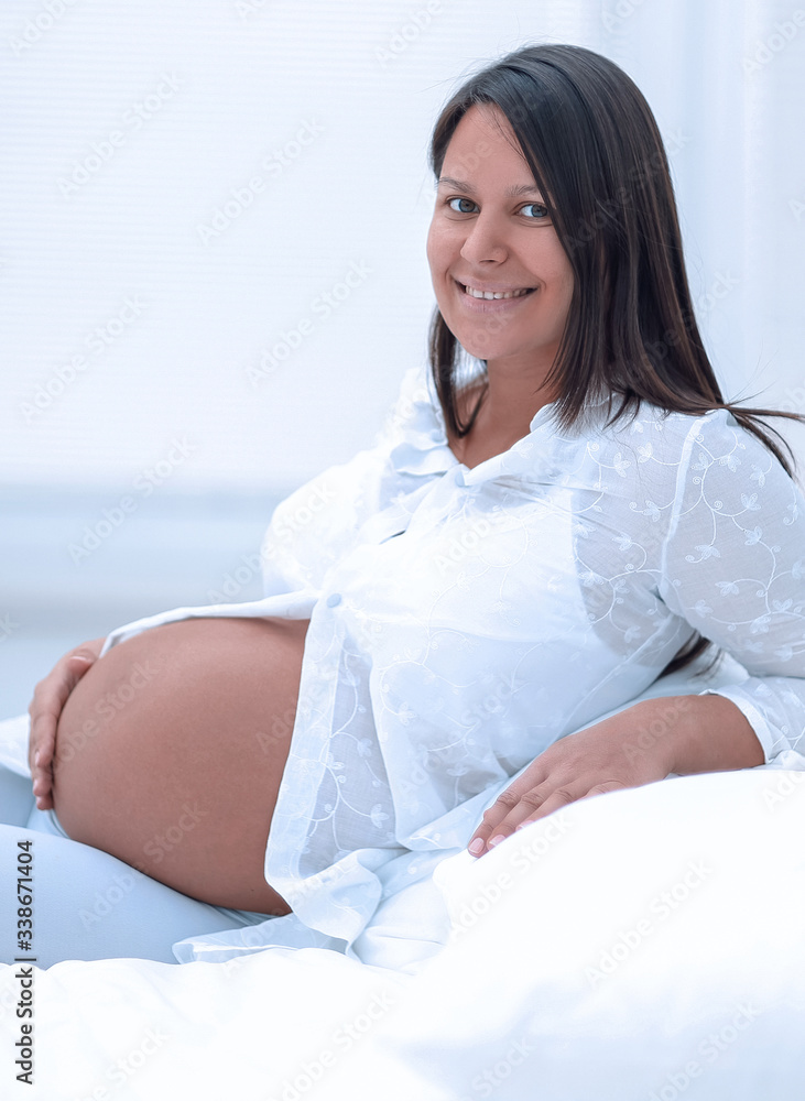 Wall mural beautiful pregnant woman sitting at sofa and keeping hand on belly.