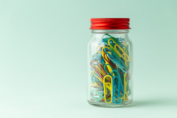 A small glass jar with a red lid full of colorful paperclips on a green background