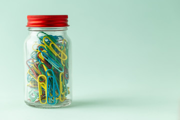 A small glass jar with a red lid full of colorful paperclips on a green background
