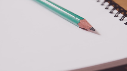 Closeup of a green pencil on a notebook. Notepad open on a blank page on the table.