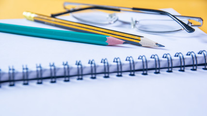 Closeup of two pencils and glasses on a notebook was open blank page, yellow desktop with soft focus.