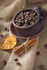 Coffee beans in a clay cup, cinnamon sticks, dried oranges on a background of burlap.