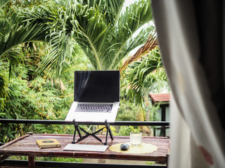 laptop of a remote digital nomad on a wooden bamboo table with notebook, mobile phone and glass in...