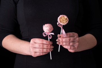 Cake pops in women's hands on a black background. Dessert in pink chocolate cream with powder and bow.