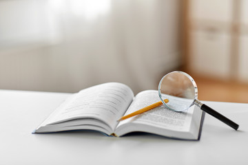 education, school and learning concept - book with magnifier and pencil on table at home