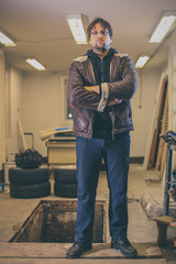 Tough young man with a beard and rock and  roll vibe standing in a garage and posing. Visible car service trench and old tires in the background. Man with arms crossed.