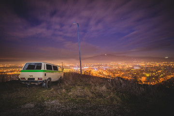 Night panorama of a city of Maribor with a van parked on top of the hill. Urban escape with a retro van, overlooking the city at night. - Powered by Adobe