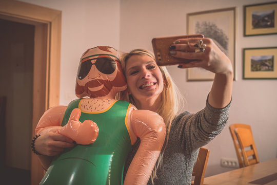 A smiling young blonde woman making a selfie wih a smartphone with an inflatable male doll. Party environment inside with a funny game and inflatable doll.
