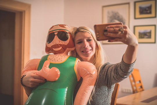 A smiling young blonde woman making a selfie wih a smartphone with an inflatable male doll. Party environment inside with a funny game and inflatable doll.