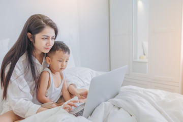 Single mom start working day on a laptop sitting on the bed with her son in the morning. Working from home concept