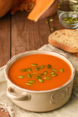 Delicious pumpkin soup in bowl on wooden table. Carrot organic vegetarian bread, seeds and slice of ripe pumpkin. Menu, recipe