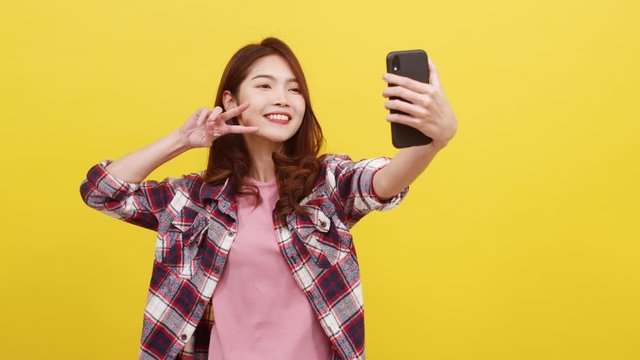 Smiling adorable Asian female making selfie photo on smartphone with positive expression in casual clothing and looking at camera over yellow background. Happy adorable glad woman rejoices success.