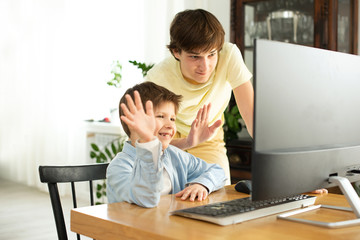 Smiling boy and teenager chatting online and waving at the computer screen. Quarantine and self-isolation due to coronavirus.