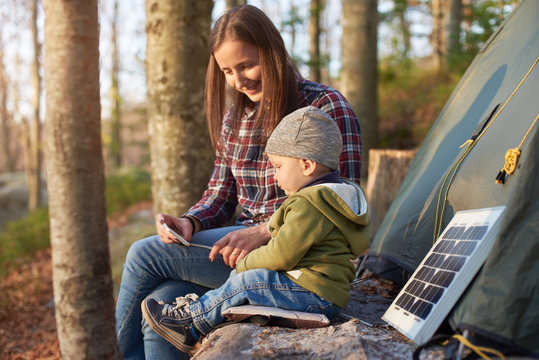 Mother With Son On Camping Vacation Looking At A Mobile Phone While Enjoying The Fresh Air. Family Sit On A Log Near The Tent And Tourist Solar Panel In The Forest On A Sunny Day.