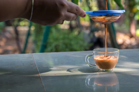a hand pouring steamy masala tea through plastic strainer into a glass cup