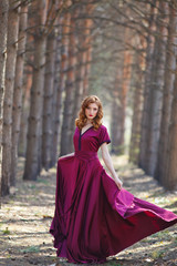 red-haired girl in a burgundy dress walks in the forest