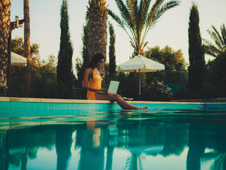 remote online working digital nomad women from the side with long hair and laptop sitting at a sunny blue water pool with plam trees in the background - obrazy, fototapety, plakaty