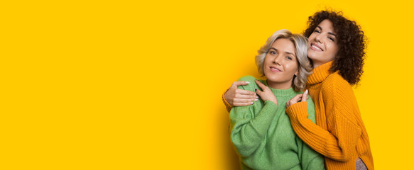 Charming girls with curly hair embracing on a yellow wall with free space while looking at camera and smile