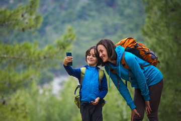 Naklejka premium A child with a backpack takes a selfie on a smartphone with mom on the background of a mountain forest.