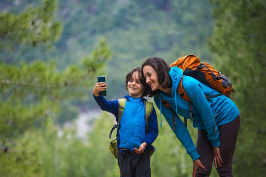 A child with a backpack takes a selfie on a smartphone with mom on the background of a mountain forest.