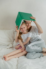 Happy young mother with daughter are reading a book in the light bedroom. They hold the book over their heads like a roof. Family relationships and home leisure. Good mood. Vertical photo