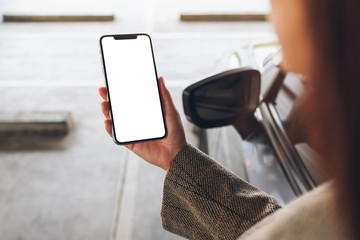 Mockup image of a woman holding and using mobile phone with blank white screen next to the car in...
