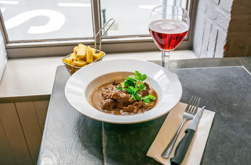Beef stew or goulash in a delicious rich brown gravy on a white plate with french fries and glass of drink over a window background