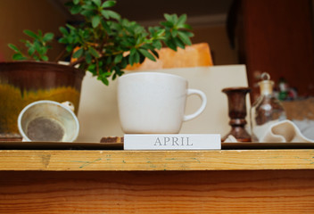 the inscription April on the background of the workplace at home during the period of quarantine and isolation. White Cup on a natural stand made of gray stone with a home green plant