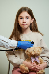 A hand of a physician holds a stethoscope hearing the heartbeat of a young female patient. A happy girl with long hair holding a Teddy bear at the doctor’s appointment.