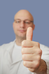 Thumbs up in focus, bald doctor with glasses in white coat out of focus, Blue background, Concept good and positeve news.