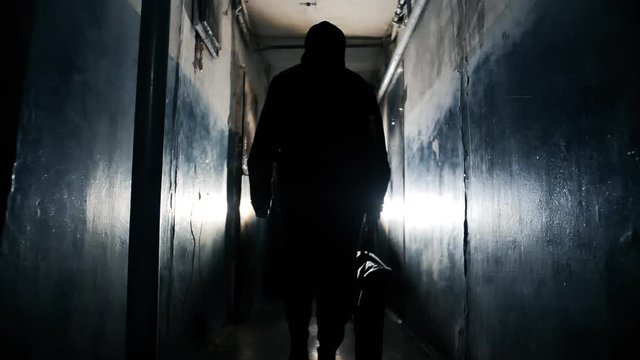 Silhouette of Unrecognizable Criminal Man Wearing Black Jacket and Carrying a Bag Walking Away From the Camera Along Hallway in Old Apartment Building Long Dark Hallway.
