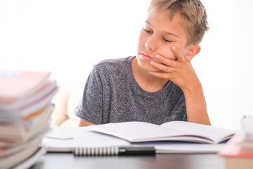 Schoolboy sitting among pile of books, textbooks, school exercise books on his desk and doing homework. Education, online learning, distance learning at home