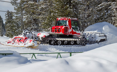 Bright red snowcat (groomer) parked in deep snowdrift after work on the ski slope against coniferous trees. Selective focus