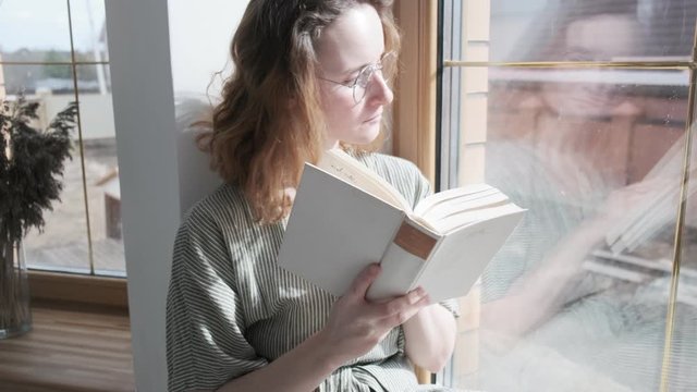 girl in dress, glasses sitting on windowsill, leafing through book. Activity, hobby during quarantine of coronavirus pandemic COVID-19, 2019-nCoV. self isolation, stay home concept. Reading poetry