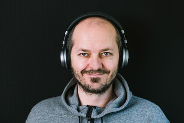 Bearded relaxed man in hoodie standing against black background with headphones, listening music, and smiling  head and shoulders
