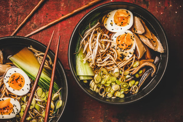 Flat-lay of traditional japanese Ramen soup with chicken meat, eggs, shiitake mushrooms in black bowls and bamboo chopsticks over dark red wooden table background, top view. Japanese cuisine concept