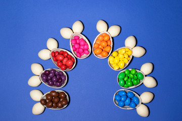 Rabbits made of chocolate eggs and colored jellies form a semicircle on a blue background. Each rabbit coincides with one color of jelly. Photo was taken by flat lay. There is a place for text.