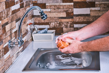 Man washing food due to coronavirus. Cleaning a tangerine to remove viruses. Metalic sink and soap to clean fruit.