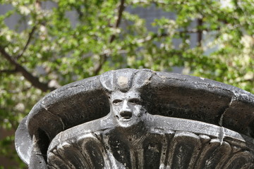 Head with face of woman or scull on the stone fountain between the green trees in the park