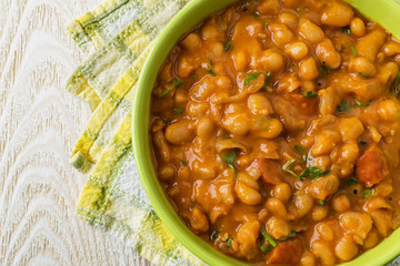 Close-up of backed beans with sausage slices in tomato sauce and parsley in a green bowl on a wooden table. Bean is source of vegetable protein and ingredient for vegetarian food.