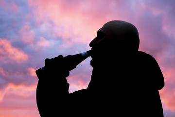 Outline of a man drinking beer from a bottle in black gloves at sunset, pink sunset clouds.