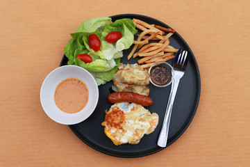 Breakfast in the black round plate on the brown floor. Fried egg with sausage, dumpling with french fries and vegetable with tomato.