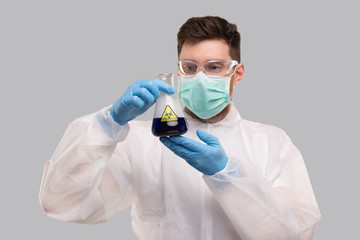 Male Laboratory Worker in Chemical Suit, Wearing Medical Mask and Glasses Watching Flask with Blue Liquid Biohazard Sign. Science, Medical, Virus Concept. Blue Liquid