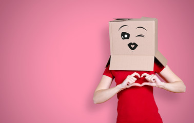Woman with cardboard box on her head and a look of love on her face, twinkling with an eye, forming a heart with her fingers on pink background