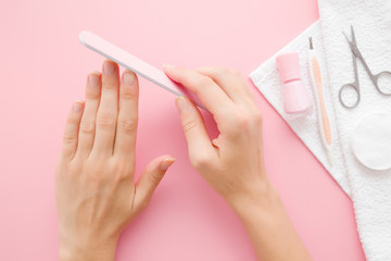Young woman hands using nail file. Simple manicure set on white towel. Pastel pink background....