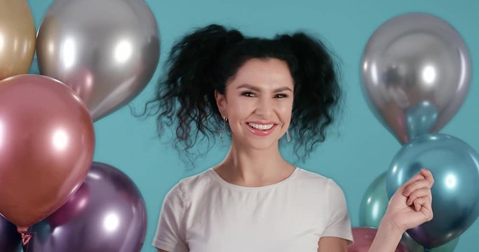 Portrait of beautiful young woman with black curly hair smiling and dancing with ballons in her hands in front of blue background shot in 4k super slow motion