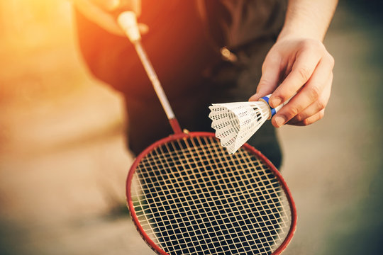 A man in dark clothing is about to throw a white shuttlecock in the air and hit it with a red badminton racket. Games in the fresh air on a Sunny day.