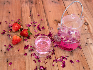 Obraz na płótnie Canvas Pink rose petals infusion tea in a small clear drinking glass with glass kettle. Wooden rustic background. 