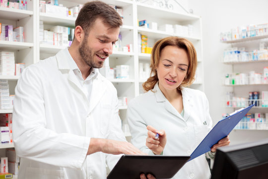 Two happy female and male pharmacists working together in a pharmacy. Young man is holding a tablet and showing something to his older colleague. Healthcare and medicine concept.
